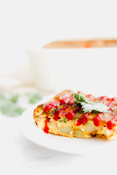 A breakfast casserole square sitting on a plate topped with hot sauce and cilantro 