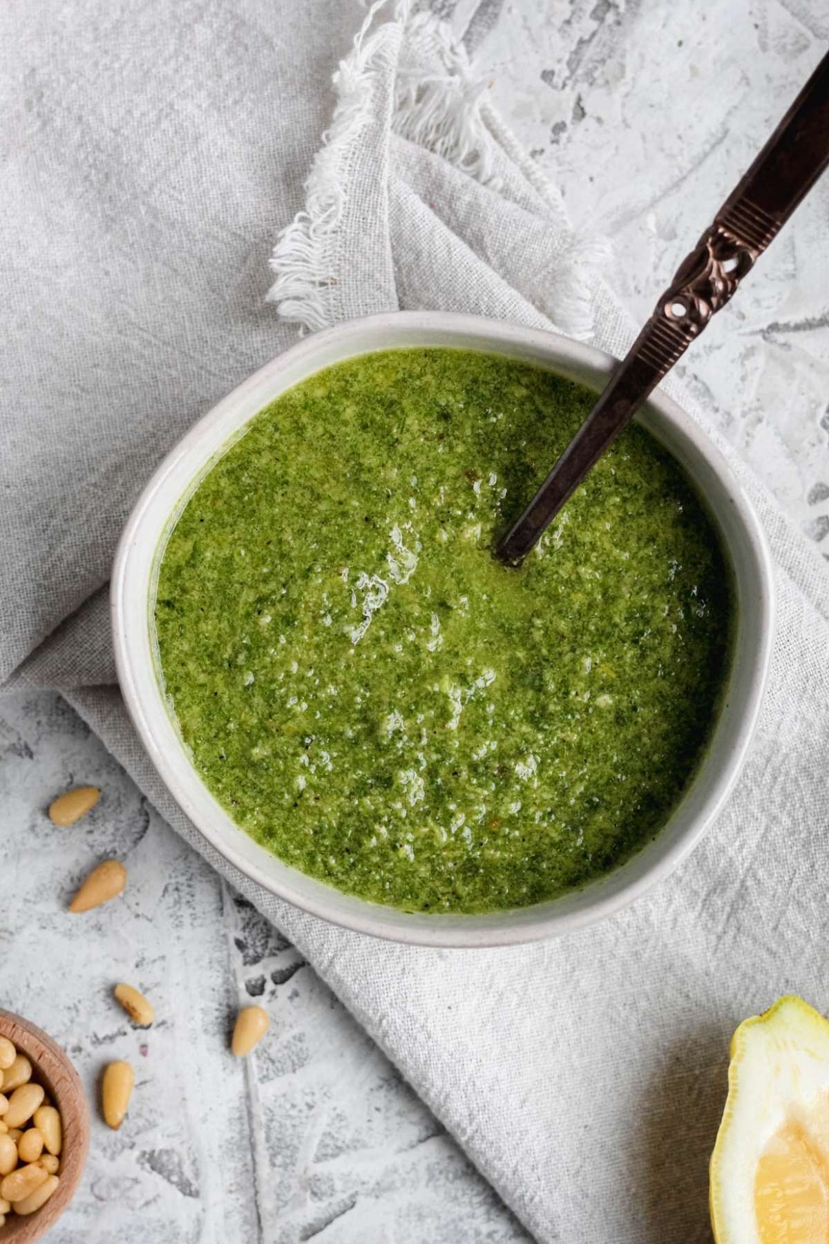 A bowl of dairy free pesto sitting on a towel with pine nuts and lemons on the sides of the bowl