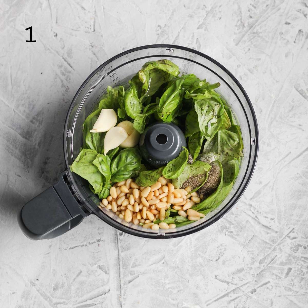a food processor filled with basil, garlic cloves, pine nuts, avocado oil, salt, and pepper