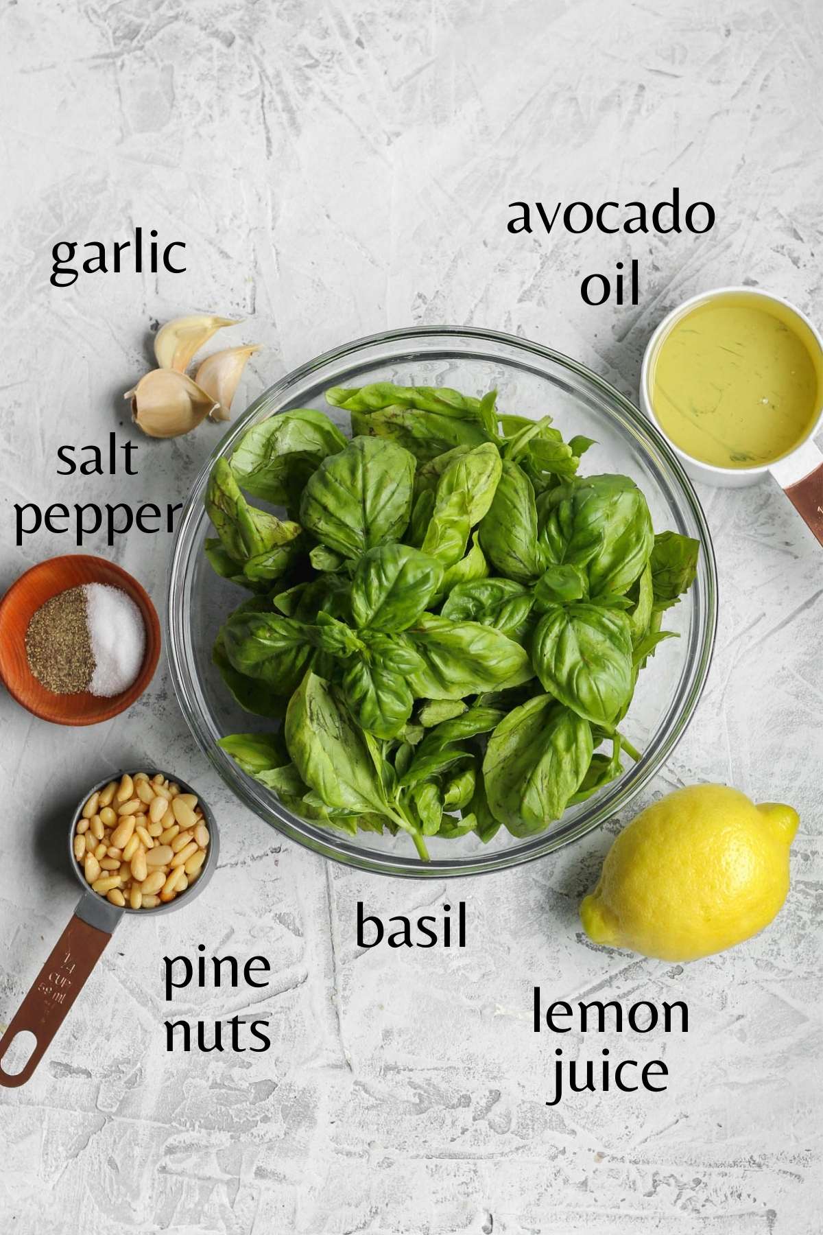 Fresh basil in a glass bowl with avocado oil in a measuring cup, garlic cloves, a lemon, pine nuts in a measuring cup, and salt and pepper in a small bowl sitting next to it