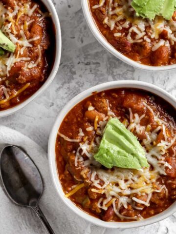 A bowl of southwestern bison chili topped with shredded cheddar cheese and avocado sitting next to a spoon.