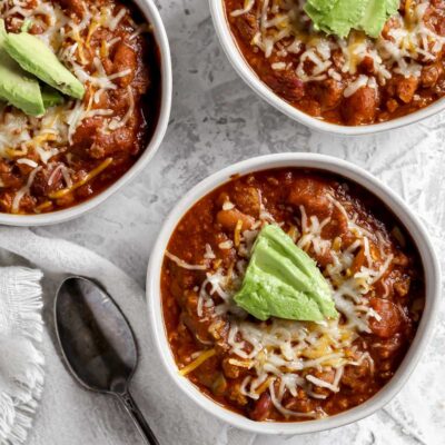 A bowl of southwestern bison chili topped with shredded cheddar cheese and avocado sitting next to a spoon.