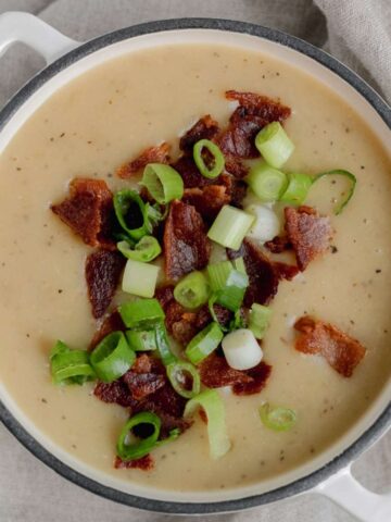 A white Dutch oven filled with gluten free potato soup that is topped with crispy bacon crumbles and fresh green onions.
