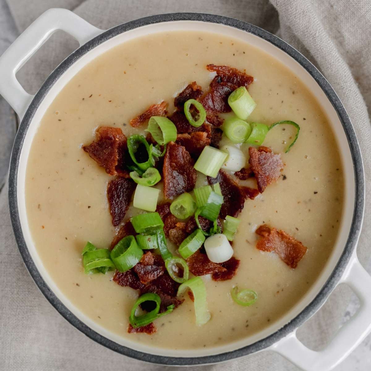 A white Dutch oven filled with gluten free potato soup that is topped with crispy bacon crumbles and fresh green onions.