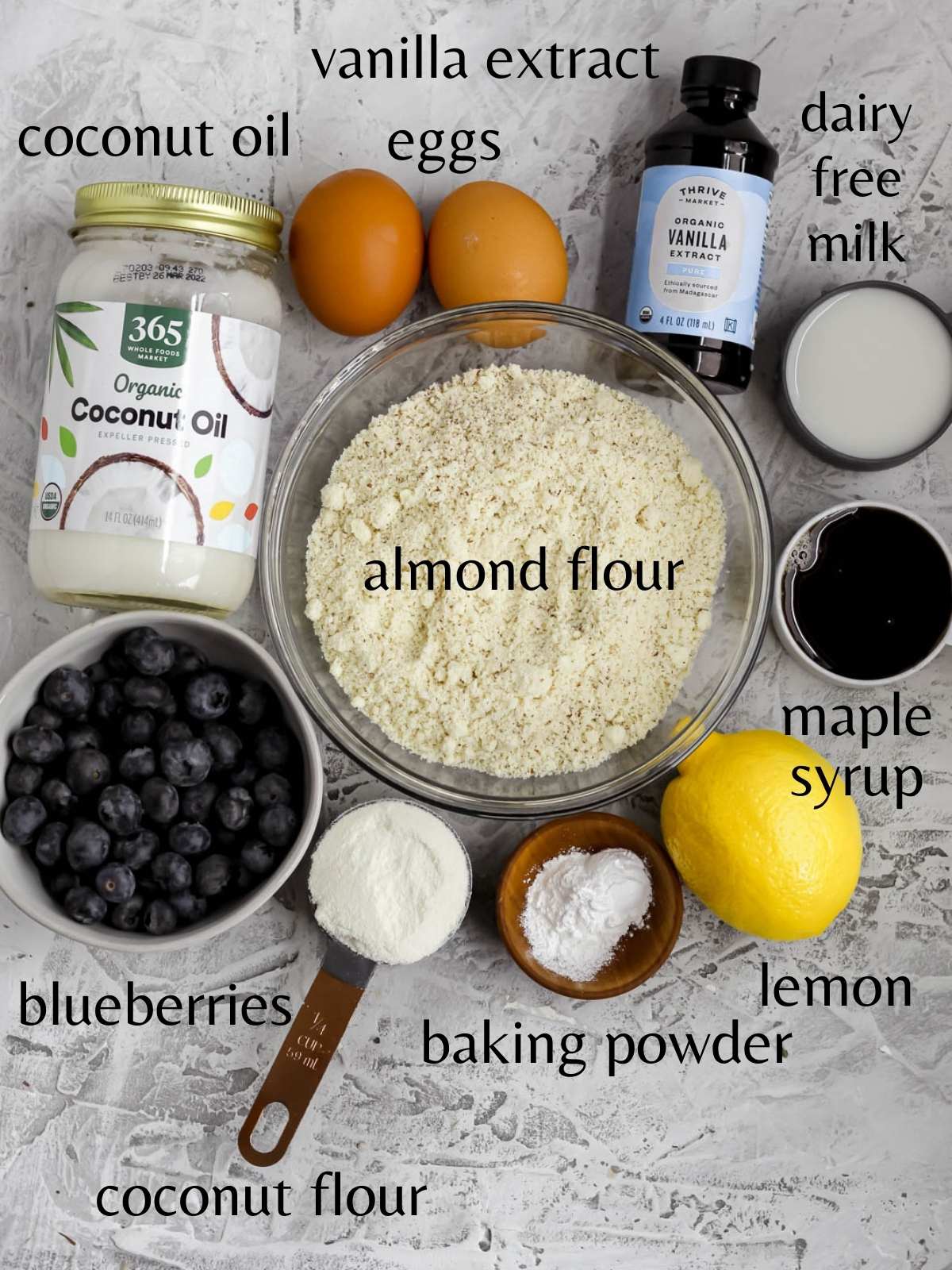 A bowl with almond flour, a small bowl with blueberries, a jar of coconut oil. 2 eggs, a bottle of vanilla extract, a small cut of dairy free milk, a small cup of maple, syrup, a lemon, a small cup with coconut flour, and a small bowl with baking powder