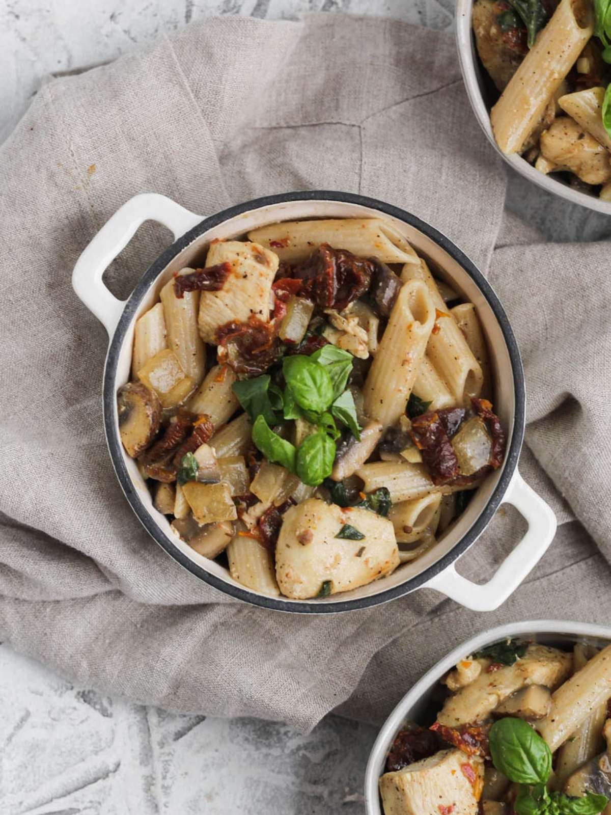 A dutch oven pot sitting on a tan towel filled with creamy sun dried tomato pasta and basil on top