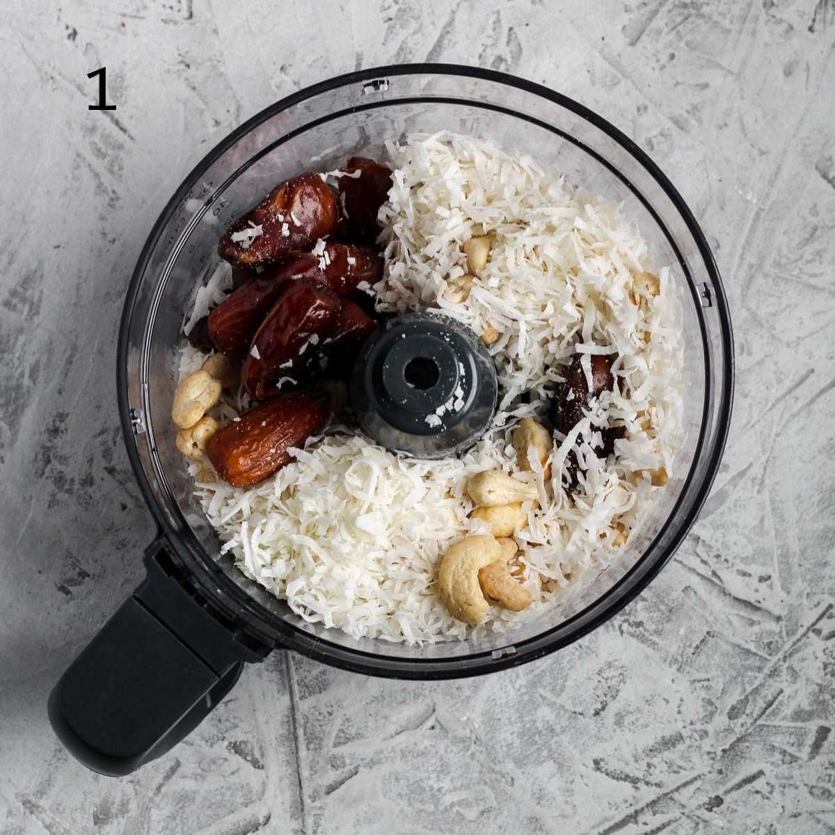 a food processor filled with pitted dates, shredded coconut, cashews, almonds, and coconut oil