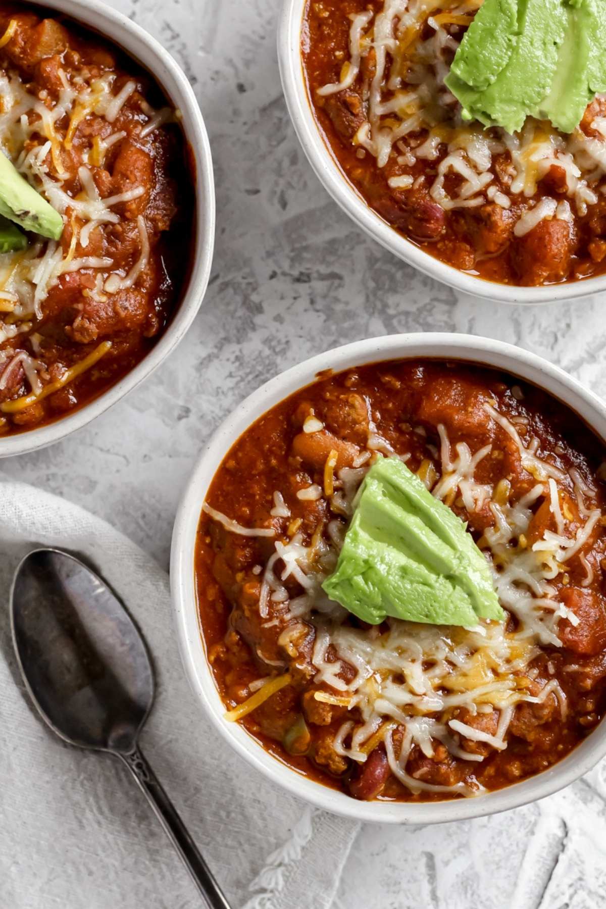 a bowl of southwestern bison chili topped with shredded cheddar cheese and avocado sitting next to a spoon 