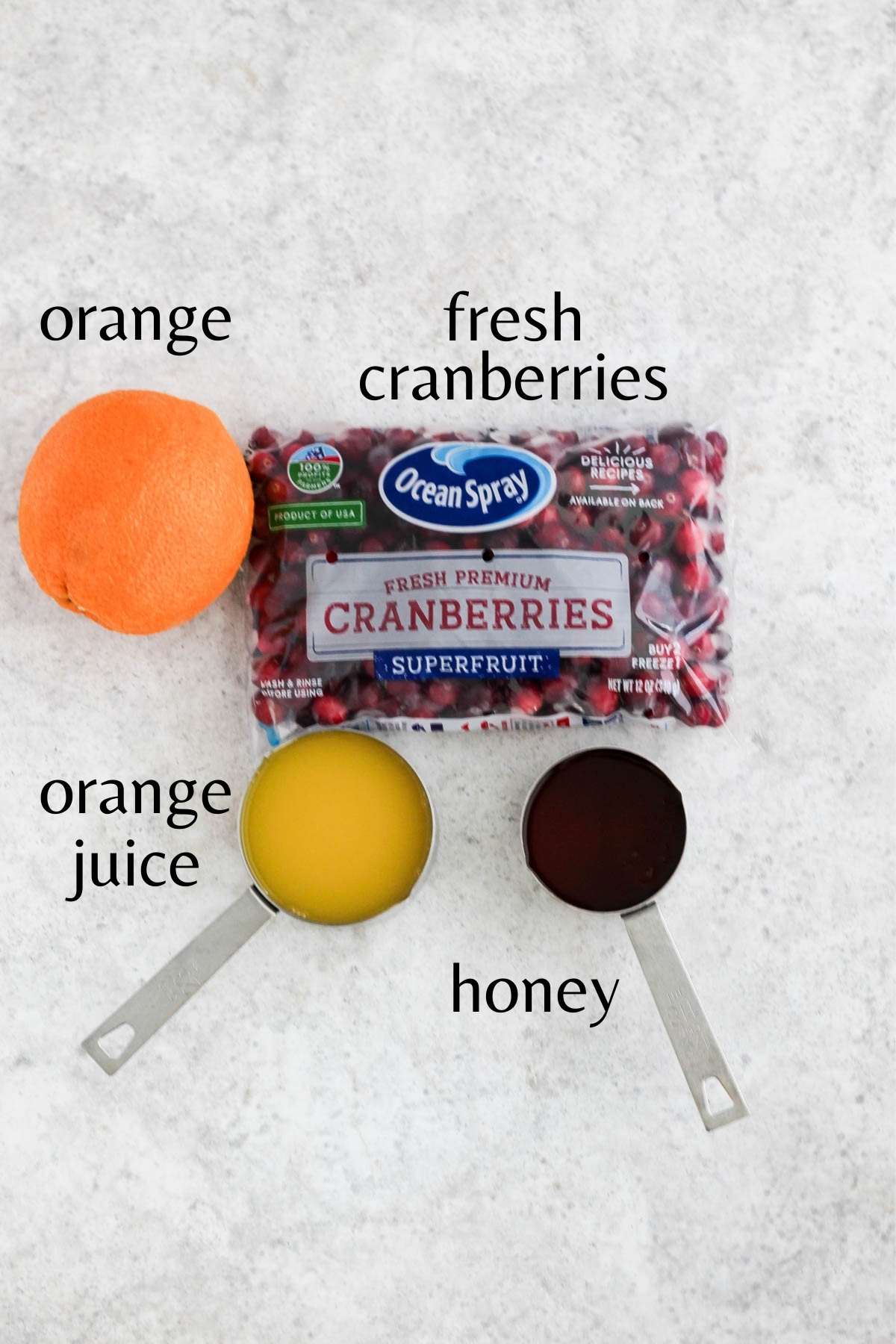 An orange, a bag of fresh cranberries,  honey in a measuring cup, orange juice in a measuring cup