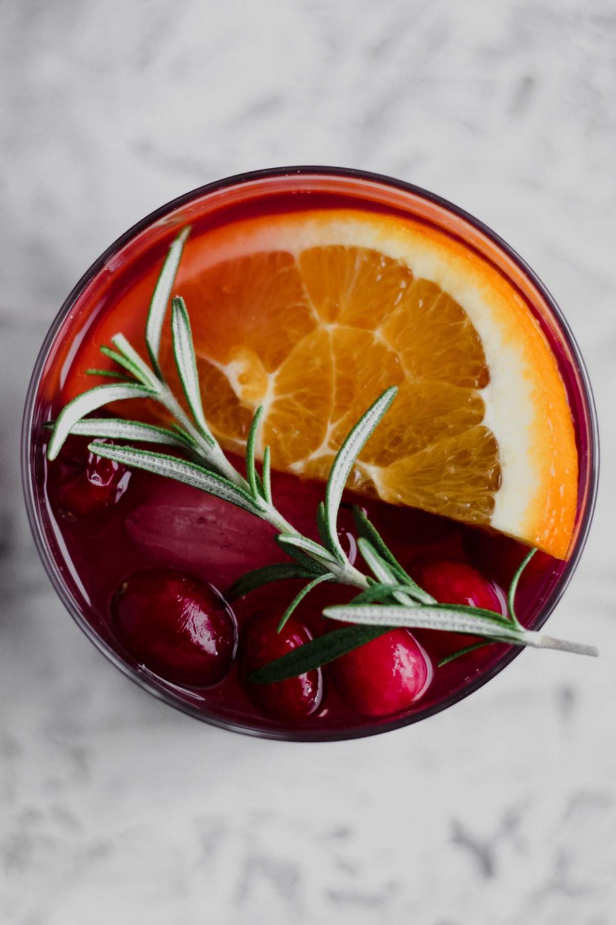 A top view of a glass filled with cranberry sangria, fresh cranberries, an orange slice, and a rosemary sprig.