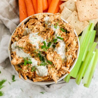 Dairy free buffalo chicken dip in a white bowl, topped with a drizzle of ranch, chives, and dill sitting on a brown cutting board with carrots, celery, and cracker also on the board.