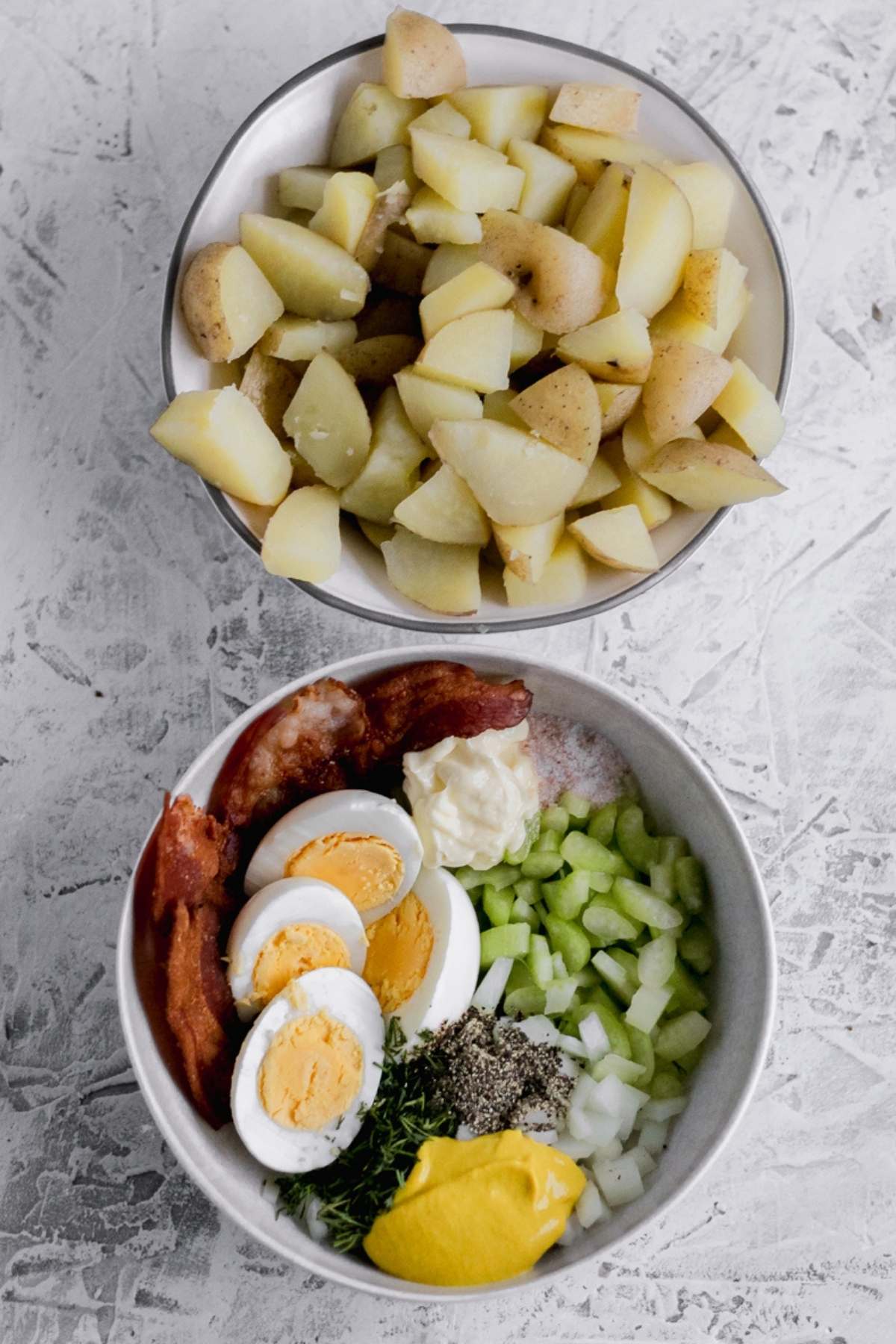A bowl of cooked potatoes and a bowl filled with cooked bacon, hard boiled eggs, mayo, seasonings, diced celery and diced onion.