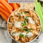 A Pinterest pin for dairy free buffalo chicken dip with a bowl of dairy free buffalo chicken dip, ranch and fresh dill drizzled on top, sitting next to carrot sticks, crackers, and celery sticks.