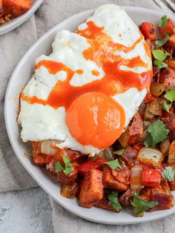 A plate filled with sweet potato hash topped with a sunny side up egg and hot sauce.