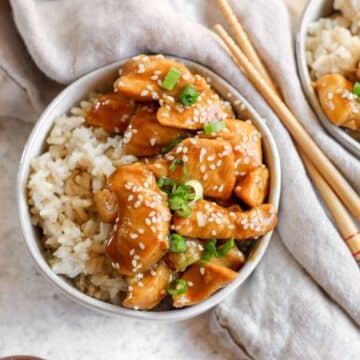 Gluten free chicken teriyaki sitting on top on rice in a round bowl with a tan tea towel and chopsticks on the side of the bowl.