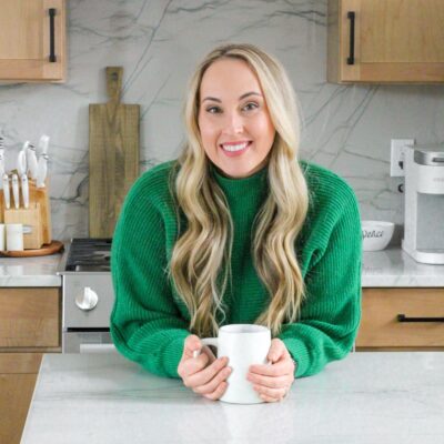 A picture of Kaleena in a green sweater holding a coffee mug leaning on the counter in a kitchen.