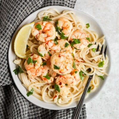 A plate filled with gluten free shrimp scampi, a fork twirled with pasta on the side of the plate, and a lemon wedge on the side of the pate all sitting on a black tea towel.