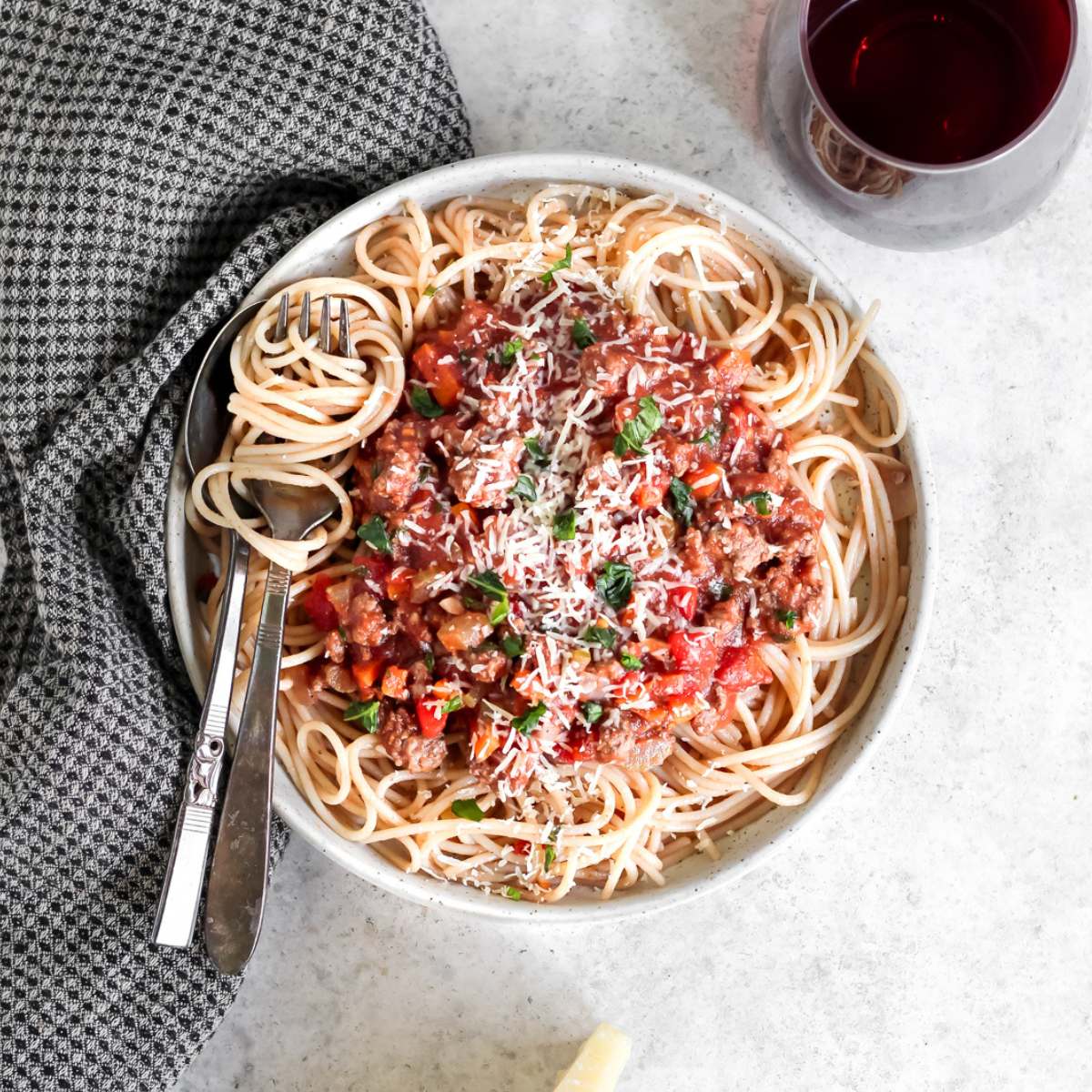A plate filled with gluten free spaghetti topped with bison bolognese, a fork with twirled spaghetti on the plate, a glass of red wine in the corner. and a black tea towel on the side of the plate.