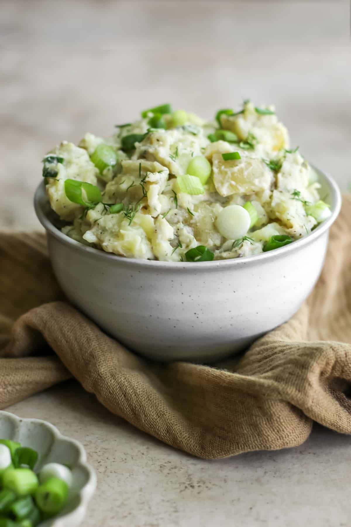 A bowl of potato salad with spring onions sitting on a tan tea towel.