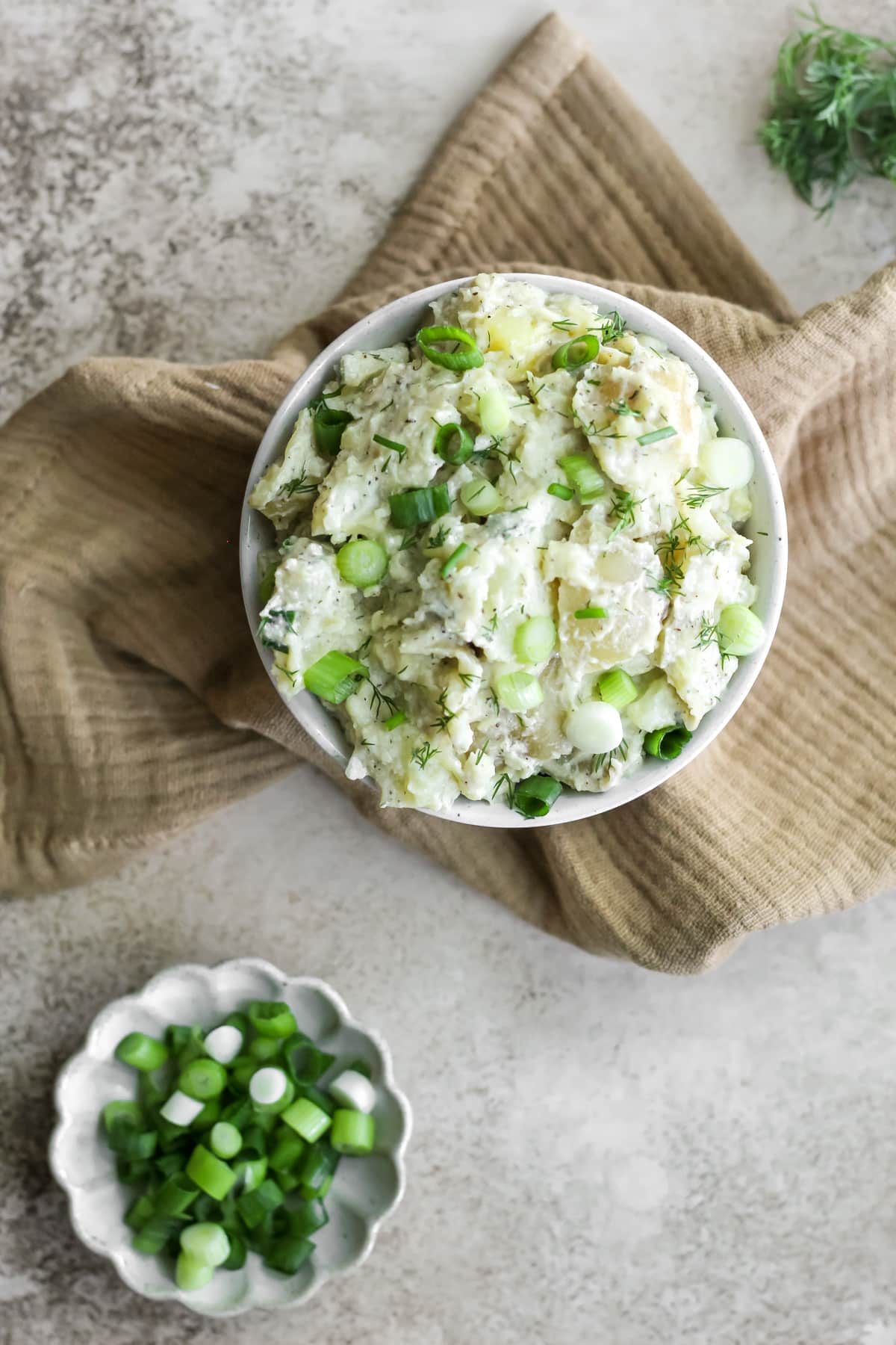 A bowl of potato salad with spring onions sitting on a tan tea towel with a little plate of diced spring onions in the bottom left corner.