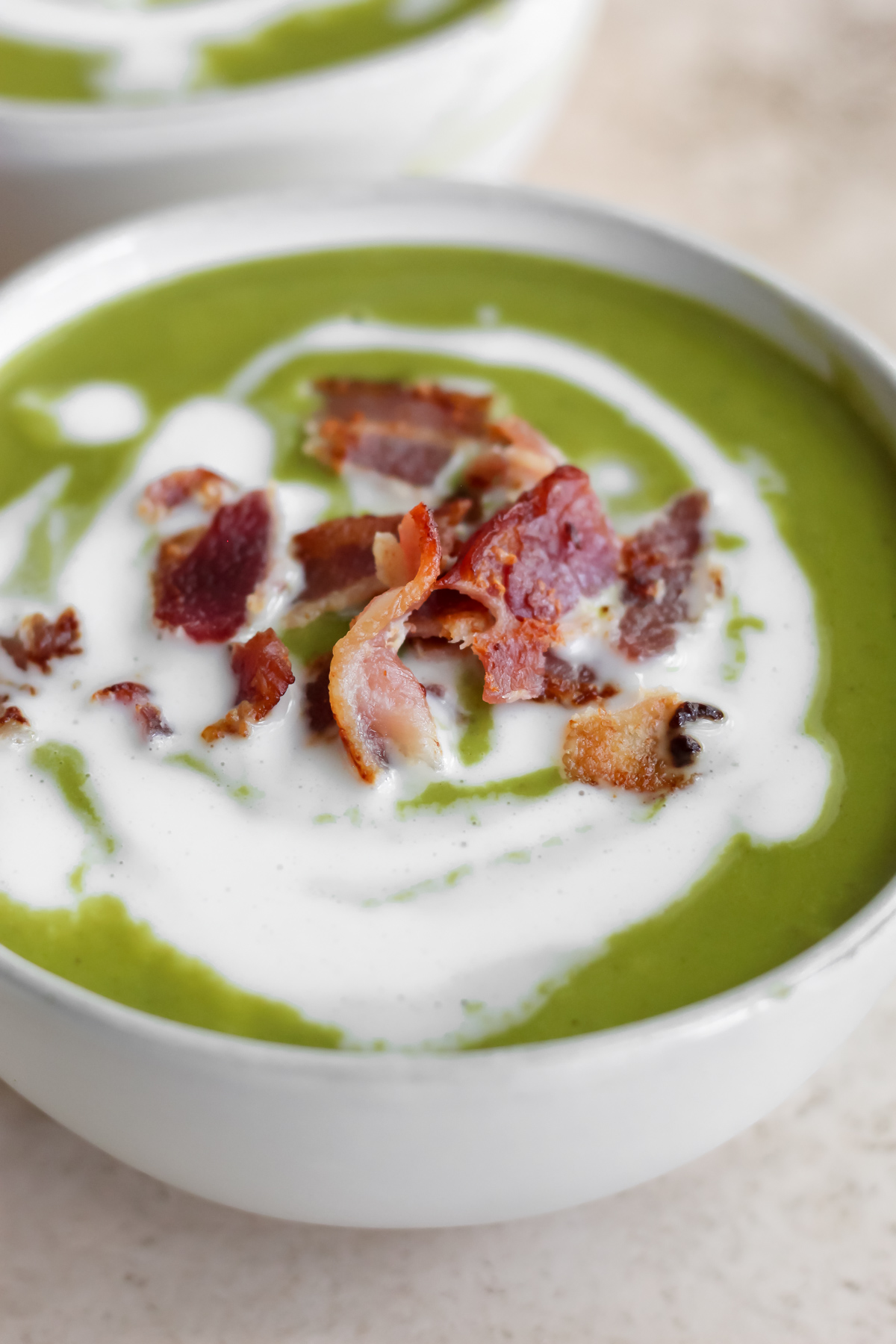 A bowl of broccolini soup with a white drizzle of coconut milk and a sprinkle of crispy bacon on top.