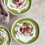 Three bowls of broccolini soup with a white drizzle of coconut milk and a sprinkle of crispy bacon on top.
