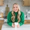 A picture of Kaleena in a green sweater leaning on a kitchen counter holding a coffee mug.