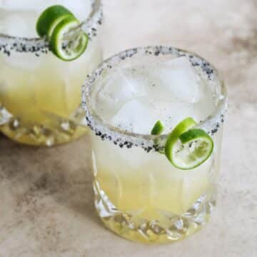 Two glasses of topo chico margaritas with curly lime twists on the side of a a cocktail glass rimmed with white and black salt.