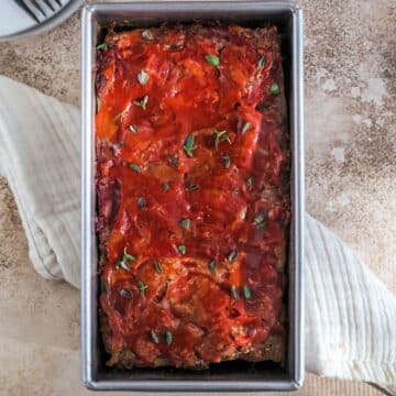 A meatloaf pan filled with Whole30 Meatloaf, sprinkled with fresh thyme on top, sitting on top of a cream colored tea towel.