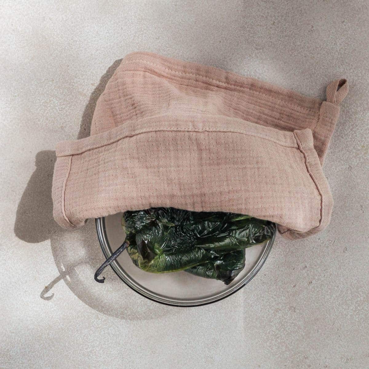 Roasted poblano peppers inside a bowl with a towel half covering the top of the bowl.